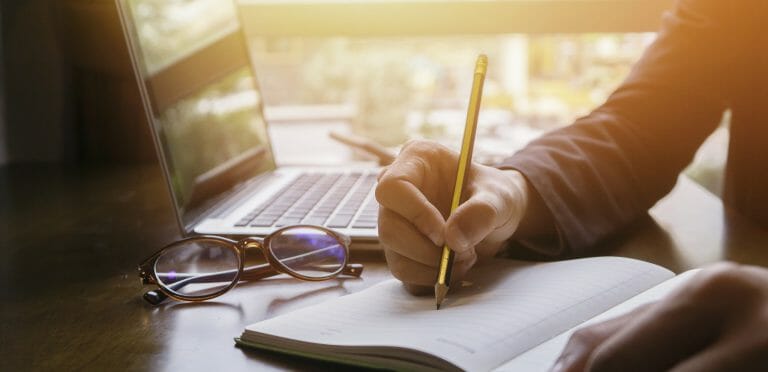 Je ziet iemand schrijven in een notitieboek met een potlood. Er staat ook een laptop open.